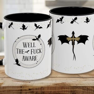 May include: White ceramic mug with a black interior and handle. The mug features a design with black dragon silhouettes and the text "WELL THE FUCK AWARE" in a circular frame. Small gold dots are scattered around the design.