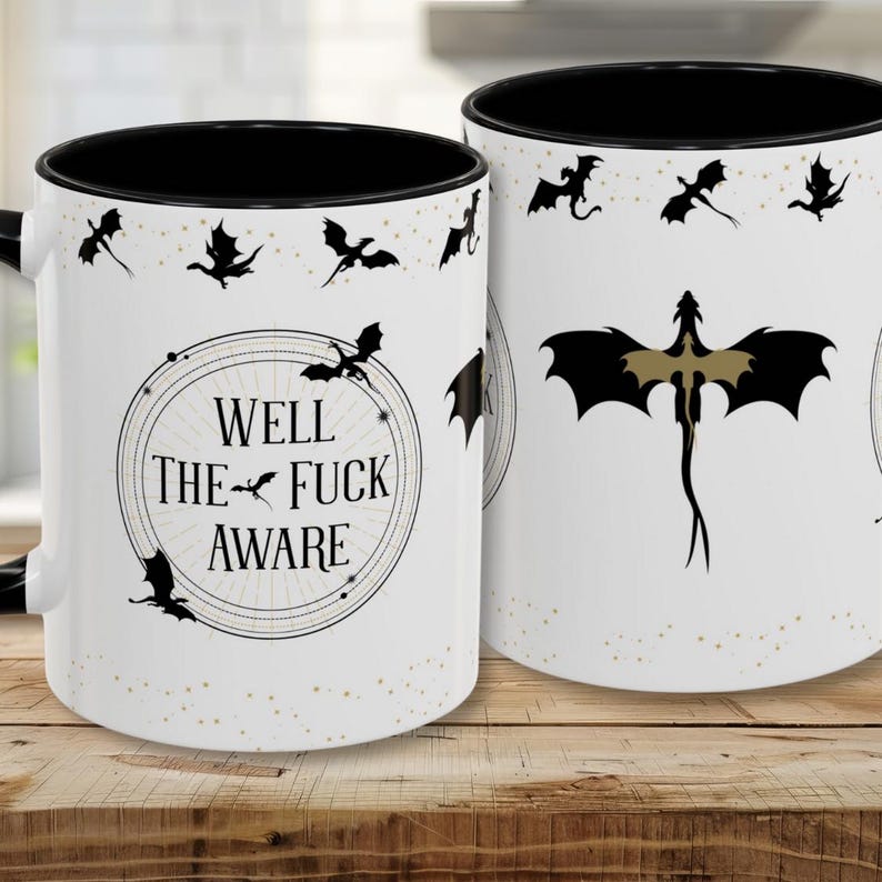 May include: White ceramic mug with a black interior and handle. The mug features a design with black dragon silhouettes and the text "WELL THE FUCK AWARE" in a circular frame. Small gold dots are scattered around the design.