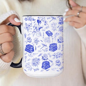 May include: White ceramic mug with a black handle and rim, featuring a blue and white floral and book design. The design includes illustrations of flowers, books, birds, and a woman reading. A decorative and functional item.