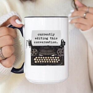 May include: White ceramic mug with a black and navy blue handle and rim. The mug features a black and silver vintage typewriter graphic with the text "currently editing this conversation."