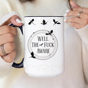 May include: White ceramic mug with a black handle and rim. The mug features the text "WELL THE FUCK AWARE" inside a circular design, with black dragon silhouettes and gold speckles.