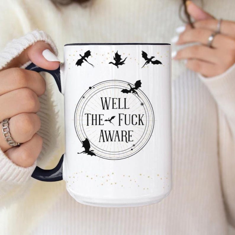 May include: White ceramic mug with a black handle and rim. The mug features the text "WELL THE FUCK AWARE" inside a circular design, with black dragon silhouettes and gold speckles.