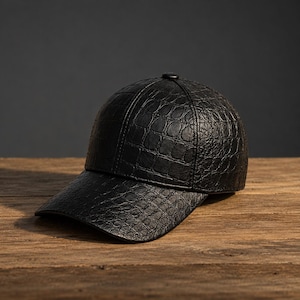 May include: A black baseball cap featuring a textured crocodile skin pattern across the entire surface. The cap has a structured crown, a curved brim, and a small button at the top centre where the panels meet, creating a uniform look.