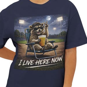 Può includere: T-shirt blu navy con un procione con un berretto da baseball, seduto su una sedia pieghevole, che tiene una birra e fa il segno della pace. Il testo "I LIVE HERE NOW" è sotto. Lo sfondo mostra un campo da baseball di notte.