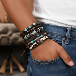 Peut inclure: Plusieurs bracelets en cuir marron ornés de perles turquoise et d'éléments argentés. L'un des bracelets est orné d'un charm en forme de tête de taureau argentée. Un autre présente un motif de peau de vache. Les bracelets sont portés sur un poignet.