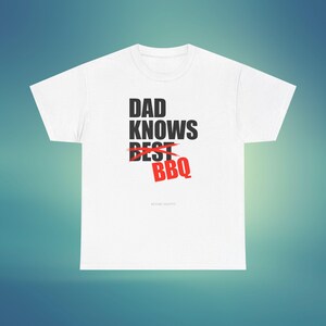 Peut inclure: T-shirt blanc avec l'inscription "DAD KNOWS BEST BBQ" en noir et rouge. Le mot "BEST" est barré. Le t-shirt a un col rond et des manches courtes. Le texte "BEYOND GRAFFITI" est en bas.