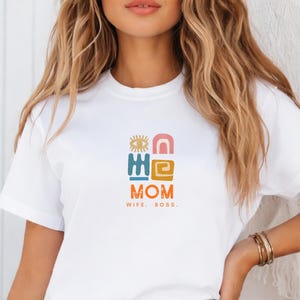 Puede incluir: Camiseta blanca con un gráfico colorido que dice "MOM" con formas abstractas y el texto "WIFE. ROSS." debajo. La camiseta se combina con unos vaqueros azules.