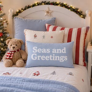 May include: A light blue rectangular pillow with the white text Seas and Greetings, placed on a bed with a white quilt featuring embroidered holiday motifs, a red and white striped pillow, a cream pillow with small icons, and a brown teddy bear.