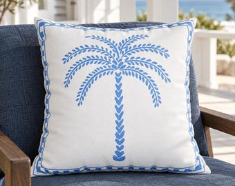 May include: A square throw pillow with a white base and a blue palm tree graphic centered on the front. The pillow features a blue decorative border with a repeating square pattern along the edges, providing a clean and symmetrical design element.