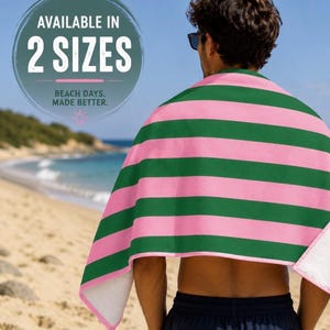Op de afbeelding: Een strandlaken met afwisselende horizontale roze en groene strepen. De handdoek is om een persoon op een zandstrand gewikkeld. De tekst "AVAILABLE IN 2 SIZES" staat in een cirkel.