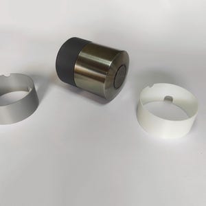 Pode incluir: Um dispositivo cilíndrico com um topo preto mate e um corpo em bronze metálico, acompanhado por dois anéis circulares separados, um com acabamento em prata mate e o outro em branco mate, cada um com um pequeno entalhe na borda superior.