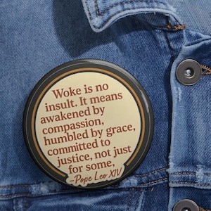May include: A round button with a black border and a tan center, featuring the quote "Woke is no insult" attributed to Pope Leo XIV. The button is set against a blue denim jacket.