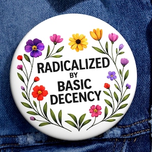 Może przedstawiać: Biała przypinka z czarnym napisem "RADICALIZED BY BASIC DECENCY", otoczona kwiatowym wieńcem z kolorowych kwiatów i zielonych liści. Przypinka jest przypięta do niebieskiej kurtki dżinsowej.