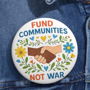 May include: A white button with the text "FUND COMMUNITIES NOT WAR" in orange and blue. The button features a graphic of two hands shaking, surrounded by flowers and hearts, against a denim jacket.