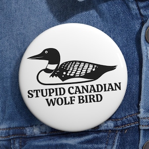 May include: A white button featuring a black and white illustration of a loon and the text "STUPID CANADIAN WOLF BIRD". The button is round and appears to be pinned to a denim jacket.
