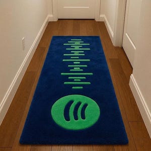 Puede incluir: Una alfombra rectangular azul con el logotipo verde de Spotify y un diseño de ondas de sonido. La alfombra está colocada sobre un suelo de madera en un pasillo, con puertas y paredes blancas al fondo.
