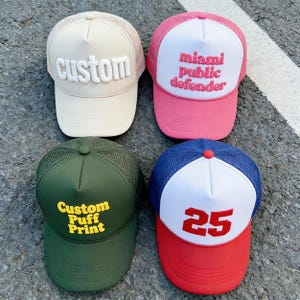 Op de afbeelding: Vier truckerpetten in verschillende kleuren. Eén is beige met "custom" in wit, een andere is roze met "miami public defender" in wit, een groene pet met "Custom Puff Print" in geel en een blauw en rode pet met het nummer "25".