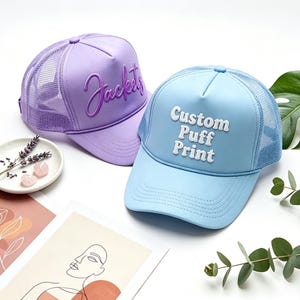 Può includere: Sono mostrati due cappellini da camionista, uno lavanda e uno azzurro. Il cappellino lavanda ha la parola "Jackets" in corsivo. Il cappellino azzurro ha le parole "Custom Puff Print". Entrambi i cappellini hanno un retro in rete.