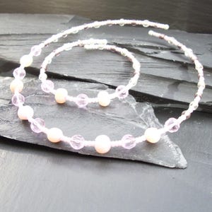 Puede incluir: Un delicado conjunto de collar y pulsera. El collar y la pulsera están hechos de pequeñas cuentas transparentes y cuentas facetadas más grandes, rosas y transparentes. Las joyas se exhiben sobre una superficie oscura y texturizada.