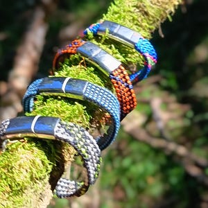Könnte beinhalten: Vier geflochtene Armbänder mit silberfarbenen magnetischen Verschlüssen. Die Armbänder weisen verschiedene Muster in Blau, Orange und Grau mit grünen Akzenten auf und sind in einer Reihe auf einer strukturierten Oberfläche angeordnet.