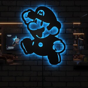 K&ouml;nnte beinhalten: Schwarze Metall-Wandkunst mit der Silhouette von Mario mit blauer LED-Hintergrundbeleuchtung. Das Kunstwerk ist an einer dunklen Backsteinmauer angebracht und erzeugt einen auff&auml;lligen Kontrast. Die ikonische Figur ist in einer klassischen Pose dargestellt.