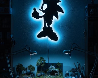 May include: A black silhouette of Sonic the Hedgehog with a blue LED backlight, mounted on a wall. The image shows a detailed miniature diorama on a desk below, with paint supplies and lamps.