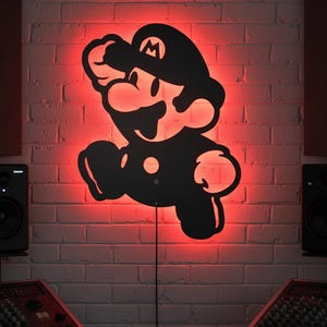 K&ouml;nnte beinhalten: Schwarze Silhouette der Videospiel-Figur Mario in Laufpose, an einer Ziegelmauer befestigt. Die Wand wird von rotem Licht beleuchtet, wodurch ein Leuchteffekt um die Figur entsteht. Ein Netzkabel erstreckt sich von der Figur.