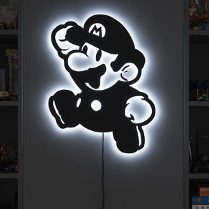 K&ouml;nnte beinhalten: Ein schwarz-wei&szlig; beleuchtetes Wandkunstwerk mit der ikonischen Mario-Figur in einer Laufpose. Das Kunstwerk hat einen hellwei&szlig;en Umriss, der einen Leuchteffekt erzeugt. Ein schwarzes Netzkabel erstreckt sich von unten.