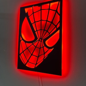 May include: A rectangular black wall-mounted sign featuring a cutout of a masked face with a web pattern. The sign is illuminated from behind with a bright red light, creating a glowing effect that highlights the intricate web design and eyes.
