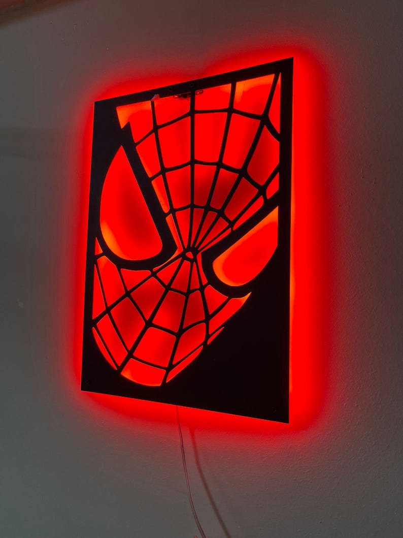 May include: A rectangular black wall-mounted sign featuring a cutout of a masked face with a web pattern. The sign is illuminated from behind with a bright red light, creating a glowing effect that highlights the intricate web design and eyes.