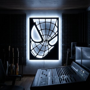 May include: A rectangular wall-mounted light fixture featuring a black cutout panel with the iconic spider web mask design. The piece is backlit with a bright white glow, creating a high-contrast silhouette of the character's eyes and web pattern.