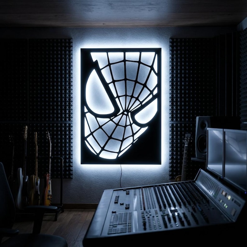 May include: A rectangular wall-mounted light fixture featuring a black cutout panel with the iconic spider web mask design. The piece is backlit with a bright white glow, creating a high-contrast silhouette of the character's eyes and web pattern.
