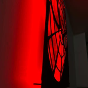 May include: A black wall-mounted panel featuring a cutout spiderweb design. The panel is illuminated from behind by a bright red light, casting a vibrant red glow onto the adjacent wall surface and highlighting the intricate web pattern.