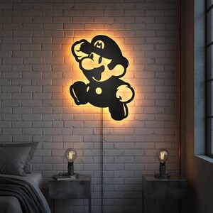 K&ouml;nnte beinhalten: Schwarze Silhouette der Videospiel-Figur Mario beim Springen, an einer wei&szlig;en Backsteinmauer befestigt. Das Kunstwerk wird von einem warmen orangenfarbenen Schein beleuchtet. Zwei kleine Nachttische mit Lampen flankieren ein Bett.