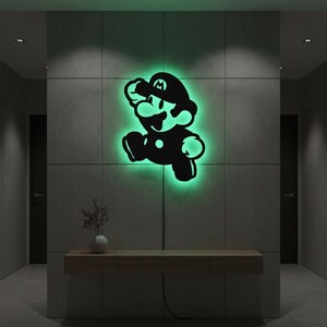 K&ouml;nnte beinhalten: Eine schwarze Silhouette der Videospiel-Figur Mario in Laufpose, beleuchtet mit gr&uuml;nem Leuchten. Das Kunstwerk ist an einer grauen gefliesten Wand in einem Flur angebracht. Unterhalb befindet sich eine Holzkonsolentisch mit einer Vase.