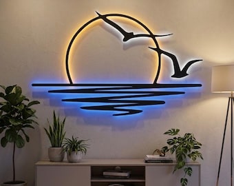 May include: A black metal wall art piece featuring a circular sun and two flying birds silhouette, illuminated by a warm yellow and cool blue LED backlight. The design includes horizontal lines representing water reflections on the wall surface.