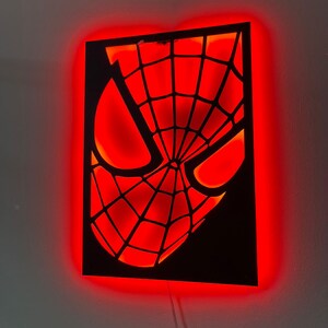 May include: A rectangular black wall-mounted light fixture featuring a cutout of a masked superhero face with a web pattern. The design is illuminated from behind by a bright red LED glow that outlines the eyes and the intricate webbed mask details.