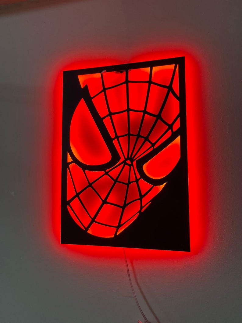 May include: A rectangular black wall-mounted light fixture featuring a cutout of a masked superhero face with a web pattern. The design is illuminated from behind by a bright red LED glow that outlines the eyes and the intricate webbed mask details.