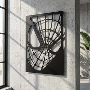 May include: A rectangular black metal wall art piece featuring a cutout design of a stylized spider mask. The design includes a prominent web pattern that covers the surface, with large, smooth eye shapes integrated into the geometric web structure.
