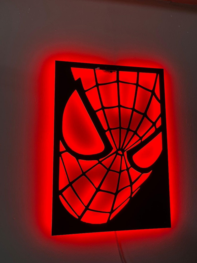 May include: A rectangular wall-mounted light fixture featuring a black cutout of a superhero mask with a web pattern. The design is illuminated from behind, casting a vibrant red glow around the edges and through the eye and web openings.
