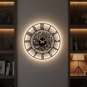Könnte beinhalten: Eine schwarze, runde Wanduhr mit arabischer Kalligraphie in der Mitte. Die Uhr hat römische Ziffern und goldfarbene Zeiger. Sie wird von einem weichen, warmen Licht beleuchtet, das einen Halo-Effekt erzeugt. Die Uhr ist an einer neutralfarbenen Wand befestigt.