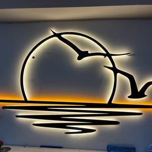 Puede incluir: Una obra de arte mural de metal retroiluminada que representa una escena de puesta de sol. El diseño presenta un sol circular, pájaros estilizados en vuelo y reflejos de agua. La pieza está iluminada con luces cálidas amarillas y naranjas.
