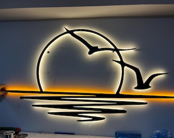 May include: A backlit metal wall art piece depicting a sunset scene. The design features a circular sun, stylized birds in flight, and water reflections. The piece is illuminated with warm yellow and orange lights.
