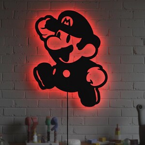 K&ouml;nnte beinhalten: Schwarze Silhouette der Videospiel-Figur Mario, mit rotem Neonlicht, das die Figur umrandet. Das Kunstwerk ist an einer wei&szlig;en Backsteinmauer angebracht. Ein schwarzes Netzkabel erstreckt sich von der Unterseite des Kunstwerks.