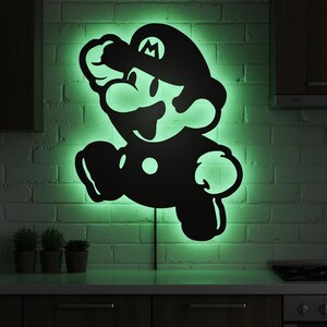 K&ouml;nnte beinhalten: Schwarze Silhouette der Videospiel-Figur Mario in Laufpose, beleuchtet von gr&uuml;nen LED-Lichtern. Das Kunstwerk ist an einer wei&szlig;en Backsteinmauer &uuml;ber einer Arbeitsplatte und Schr&auml;nken angebracht. Ein lustiges, dekoratives St&uuml;ck.