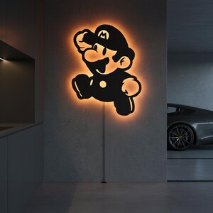 K&ouml;nnte beinhalten: Schwarze Silhouette der Videospiel-Figur Mario, beleuchtet mit einem orangefarbenen Schein. Die Wandkunst ist an einer grauen Wand angebracht. Die Figur befindet sich in einer Laufpose, mit schwarzem Hut und Latzhose.