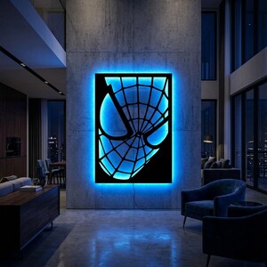 May include: A rectangular wall-mounted light fixture featuring a black cutout of a spider-webbed mask design. The piece emits a vibrant blue glow from behind the black frame, creating a silhouette effect against the concrete wall surface.