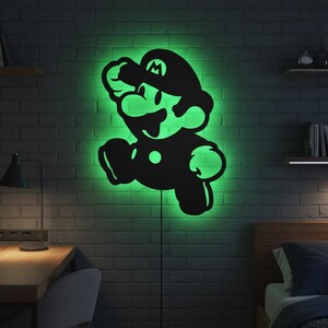 K&ouml;nnte beinhalten: Schwarze Mario-Silhouette als Wandkunst mit gr&uuml;nem Neonlicht. Die ikonische Videospiel-Figur ist in einer Sprungpose dargestellt. Das Licht ist an einer dunklen Backsteinmauer angebracht.