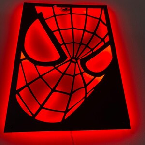 May include: A rectangular wall-mounted light fixture featuring a black cutout panel with a spiderweb mask design. The cutout is backlit with a vibrant red glow, highlighting the geometric web pattern and the large, angular eye shapes of the mask.