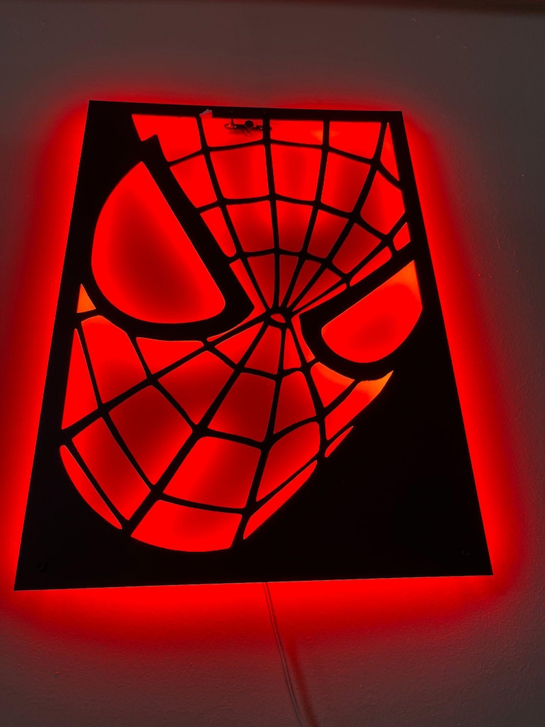 May include: A rectangular wall-mounted light fixture featuring a black cutout panel with a spiderweb mask design. The cutout is backlit with a vibrant red glow, highlighting the geometric web pattern and the large, angular eye shapes of the mask.